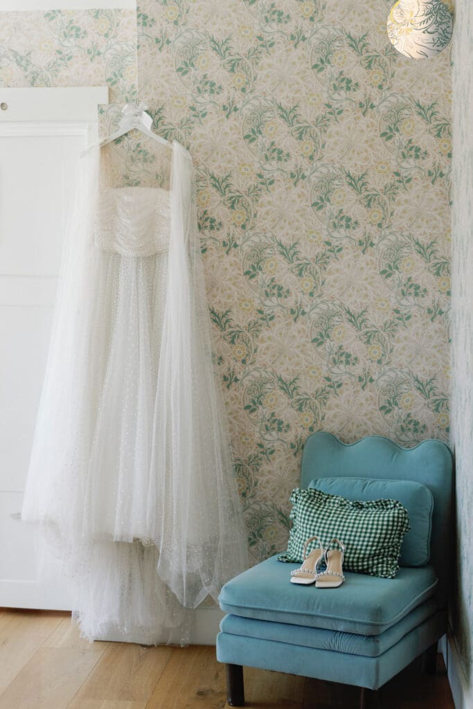 Wedding dress hanging beside a blue chair with bridal shoes in a bridal suite.